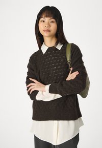 Pull en maille texturée marron, à coupe décontractée, porté par-dessus une chemise blanche à col. Le mannequin tient un sac à bandoulière vert, bras croisés.