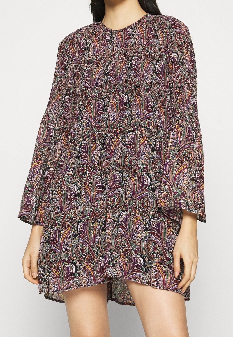 Femme portant une robe à manches longues, au-dessus du genou, avec un motif cachemire coloré et complexe dans des tons violet, orange et vert.