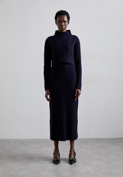 Navy ribbed knit sweater and skirt set with a high neckline, long sleeves, and a mid-calf length. Accented by black pointed-toe heels.