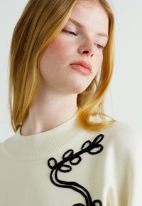 Strikkegenser i off-white med et teksturert svart blomsterbroderi på forsiden, med rund halsutringning og lange ermer.