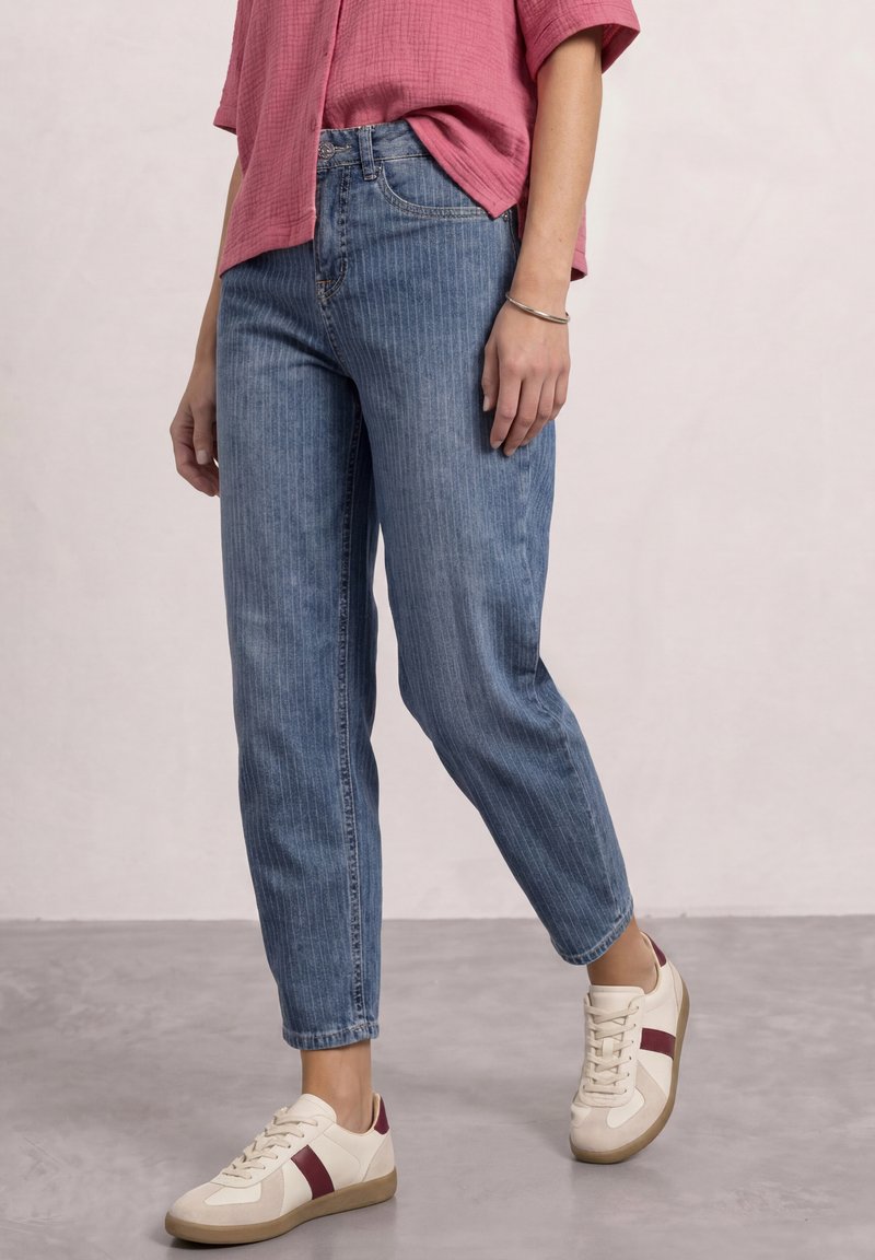 Person wearing blue tapered jeans, white sneakers with red stripes, and a pink short-sleeve shirt standing on a gray floor.