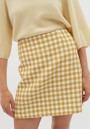 Suknja u žutom i bijelom houndstooth uzorku s teksturiranim završetkom, s visokim strukom i dužinom iznad koljena, u kombinaciji s bež majicom.