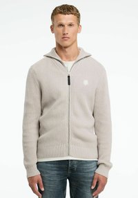 Gai bēšs ribots džemperis ar rāvējslēdzēju un augstu apkakli, ar melnu rāvējslēdzēju un mazu logotipu kreisajā pusē. Uzvilkta virs baltas blūzes.