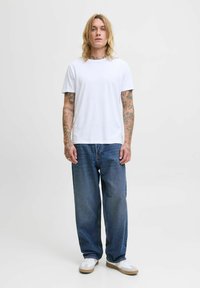 T-shirt en coton blanc associé à un jean en denim bleu à coupe ample. Il présente des déchirures légères et une silhouette décontractée, porté avec des baskets blanches.