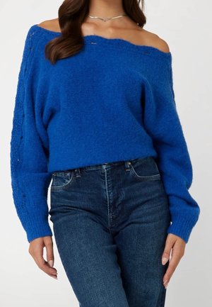 Femme portant un pull duveteux bleu royal à épaules dénudées associé à un jean taille haute bleu foncé, montrant uniquement le torse et les bras.