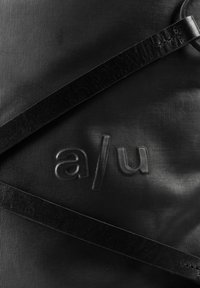 Schwarze strukturierte Tasche mit eingeprägtem "a/u" Logo und zwei schwarzen Ledergriffen, die sich diagonal kreuzen.