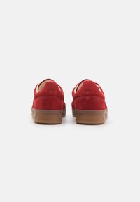Red suede sneakers with a low profile, contrasting brown rubber sole, and logo detailing on the heel. Minimalistic design with rounded shape.