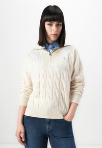 Femme avec des cheveux noirs raides portant un pull en laine crème à tresses sur une chemise bleue à col et un jean bleu, se tenant devant un fond blanc.