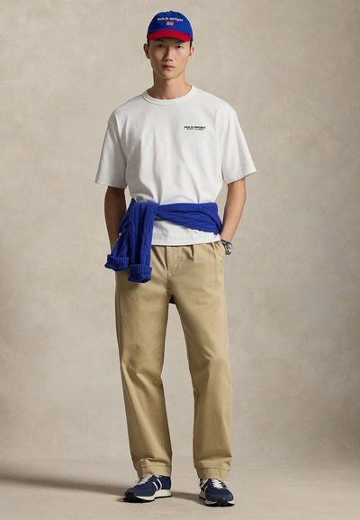 Λευκό μπλουζάκι με μαύρο λογότυπο, μπλε πουλόβερ δεμένο στη μέση, μπεζ παντελόνι και μπλε/λευκά αθλητικά παπούτσια. Αξεσουάρ περιλαμβάνουν μπλε/κόκκινο καπέλο.