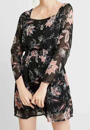 Frau trägt ein schwarzes, langärmliges Kleid mit Blumendruck in Pink und Weiß, tailliert und mit halbtransparentem Stoff an Ärmeln und Rock.