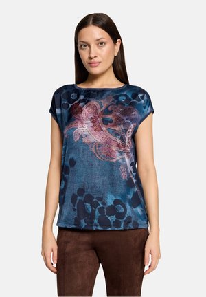 Femme aux longs cheveux foncés portant un haut bleu à manches courtes avec des motifs floraux roses et un pantalon marron, debout devant un fond uni.