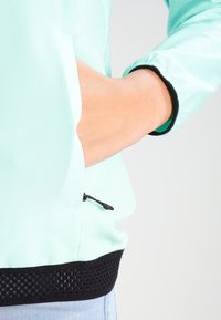 Rompevientos de color aqua claro con un bolsillo con cremallera, dobladillo de malla negra y puños elásticos. Textura suave con un ajuste relajado y un diseño minimalista.