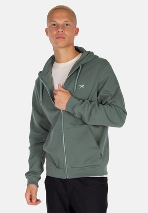 Grüne Zip-up- Kapuzenjacke mit einer Vorderseite-Tasche und verstellbarem Kapuzenzug. Hergestellt aus weichem Stoff, mit einem kleinen Logo auf der Brust.