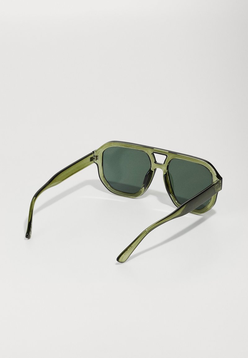 Lunettes de soleil teintées de vert avec une monture rectangulaire, présentant des branches transparentes olive et des verres gris foncé. Détails de design géométrique.