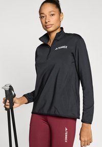 Fekete, hosszú ujjú pulóver félig cipzáros gallérral, fehér logóval a mellkason. A figura túrabotokkal a kezében. Bordó leggings látható.