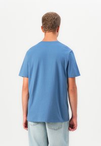 Tânăr cu păr creț scurt, purtând un tricou simplu albastru și blugi deschiși, stând cu fața spre un fundal alb.
