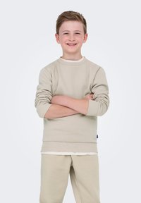 Beige katoenen sweatshirt met geribbelde manchetten en onderrand, gecombineerd met bijpassende beige joggingbroek. Kind staat met gekruiste armen en lacht.