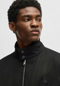 Schwarze Jacke mit Reißverschluss, hohem Kragen, zwei Knöpfen und gesticktem Logo auf der linken Brust. Glattes, leichtes Material.