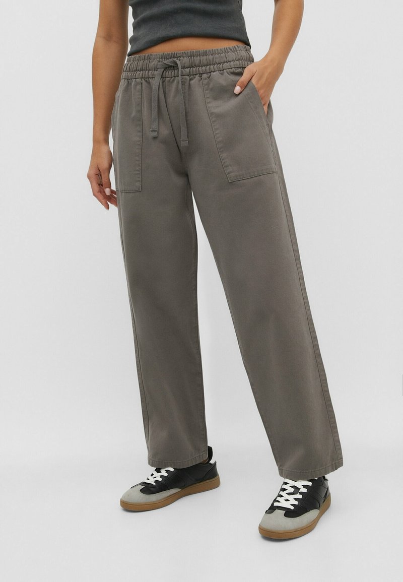 Henkilö, joka on pukeutunut avariin harmaisiin housuihin, joissa on nyöri vyötäröllä ja läpivientitaskut, sekä mustiin ja harmaisiin lenkkareihin, joissa on valkoiset nauhat.