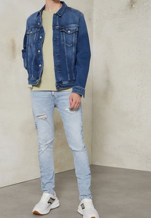 Homme portant une veste en jean bleue, un t-shirt vert clair, un jean skinny bleu clair déchiré et des baskets blanches, debout à l'intérieur.