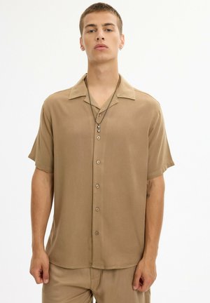 Joven con camisa beige de manga corta abotonada y pantalones a juego, con un collar de cadena y un pequeño pendiente, posando sobre fondo blanco.