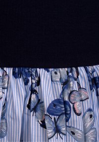 Sininen neulekangas kerätyn hameen päällä, jossa on vaaleansinisiä ja valkoisia raitoja sekä sinisiä perhosia mustilla ja vaaleanpunaisilla yksityiskohdilla.