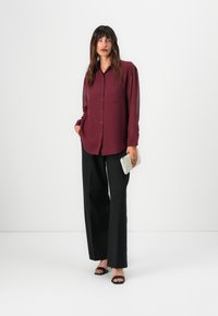 Camicia bordeaux con bottoni e vestibilità rilassata, abbinata a pantaloni neri a gamba larga. La modella tiene una pochette grigia texture, indossando sandali neri.