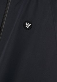 Materialul negru al jachetei de tip windbreaker are o textură netedă, prezentând un logo brodat rotund în alb. Are un fermoar la închidere în față.