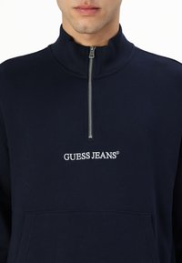 Marineblå sweatshirt med høj krave, halv lynlås, frontlomme og broderet "GUESS JEANS" logo i hvid. Blød tekstur.