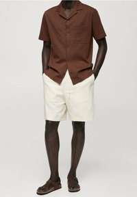 Hombre con camisa marrón de manga corta abotonada, pantalones cortos color crema y sandalias marrón oscuro con hebillas, de pie con las manos en los bolsillos.