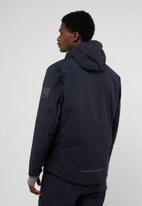 Marineblaue Regenjacke mit Kapuze, ausgestattet mit einer glatten Oberfläche, verstellbaren Manschetten und einem reflektierenden Streifen im unteren Rückenbereich. Logo auf der Schulter.