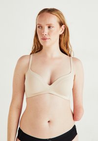 Woman with light skin and red hair wearing a beige bra and black underwear, visible left arm and right arm below elbow missing, neutral background.