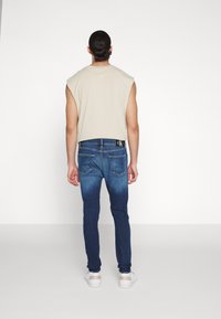 Camisa sin mangas color beige, jeans de mezclilla con efecto de desvanecimiento y zapatillas blancas. Los jeans tienen un logo en la cintura y en los bolsillos traseros.