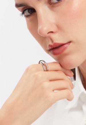 Primer plano de una persona apoyando la barbilla en la mano, llevando un anillo de doble banda plateado, con maquillaje neutro y una camisa blanca con cuello.
