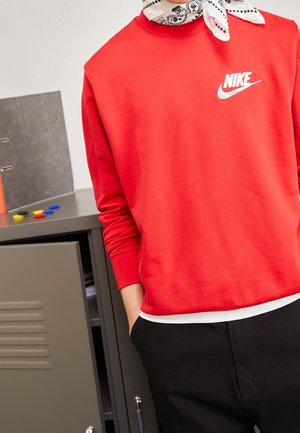 Person iført en rød Nike sweatshirt og hvid bandana om halsen, står ved siden af et åbent gråt skab med farverige markører på toppen.