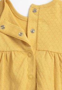 Vêtement en coton jaune avec tissu texturé, doté de pressions au niveau de l'encolure et de la taille. Présente un froncé au niveau de la taille.