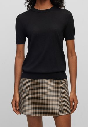 Femme portant un pull à manches courtes noir et une mini-jupe à carreaux marron et noir, debout devant un fond uni.