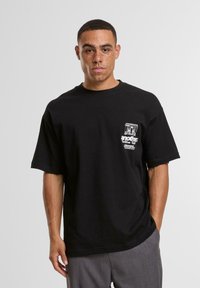 Tricou negru din bumbac, cu croi lejer, mâneci scurte și imprimeu grafic alb pe partea stângă a pieptului. Textură netedă, design casual.