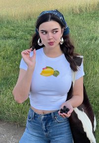 Balta trumpa viršutinė dalis su citrinos grafika, mėlynos džinsai, laikantis juodųjų serbentų, įsikūrusi žolėtoje pievoje su ruda ir balta karvės raštais dekoruota antklode.