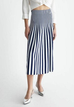 Kvinde iført midi nederdel med navyblå og hvide lodrette striber, hvid cropped bluse og sølvfarvede spidse slingback hæle.