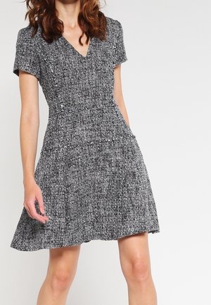 Woman wearing short-sleeve, black-and-white textured A-line dress with V-neck and fitted waist, standing against a plain light background.