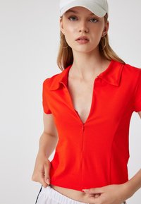 Rode polo met korte mouwen en een rits, met een slim fit ontwerp en zachte stof. Model past de zoom aan en draagt een witte pet.