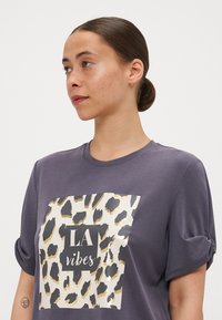 Hallitus t-särk, millel on veerandkäised, suur leopardi prindi graafika ja tekst "LA vibes" mustas ja kuldsetes toonides. Pehme tekstuur.