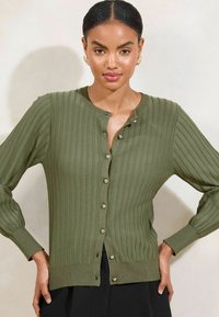Olivgrüner gerippter Cardigan mit goldenen Knöpfen, Rundhalsausschnitt und langen Ärmeln. Hat einen geraden Saum und seitliche Schlitze.
