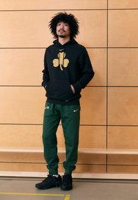 Sweatshirt preta com capuz e gráfico de trevo dourado, combinada com calças de treino verdes com logótipo branco da Nike. Modelo usa ténis pretos.