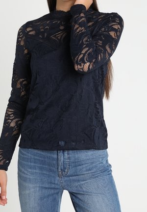 Top lungo a maniche lunghe in pizzo blu navy con motivi floreali, caratterizzato da un colletto alto e maniche trasparenti, abbinato a jeans azzurri chiari.