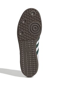 Vit sneakersula med en brun gummiyttersula, som har ett strukturerat mönster, cirkulära grepp och en logotyp nära hälen. Tre gröna ränder.