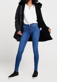 Manteau noir avec bordure en fourrure, chemise blanche à boutons, jean skinny bleu et chaussures noires sans lacets. La tenue présente un style décontracté et ajusté.