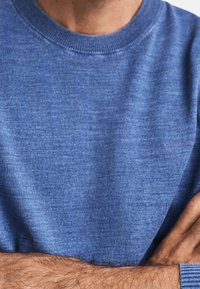 Man som bär en blå rundhalsad tröja med korsade armar, som visar den texturerade stickade tyget och en del av hans nacke och underarmar.