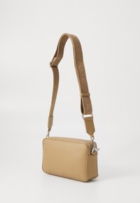 Sac bandoulière tan avec une finition texturée, fermeture éclair et sangle ajustable ornée de logos Calvin Klein en relief.